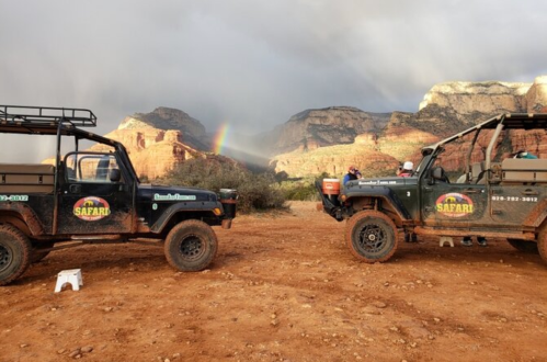 Two safari vehicles parked on a dirt path with mountains and a rainbow in the background.