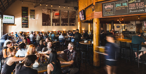 A bustling restaurant interior with patrons enjoying food and drinks at tables, featuring wooden beams and vibrant decor.
