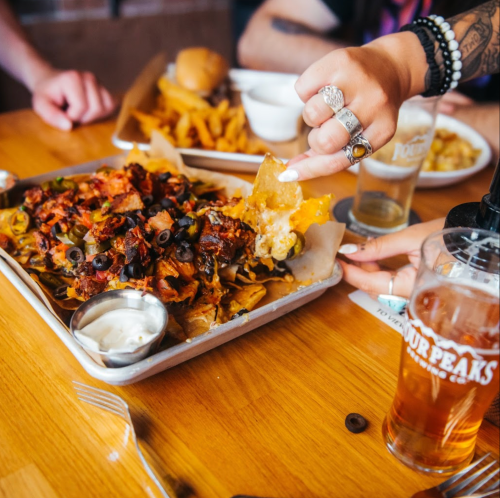 A hand reaches for a loaded nacho platter with toppings, accompanied by a glass of beer and other food on the table.