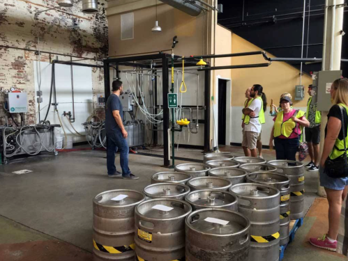 A group of people in safety vests observes a brewing area with kegs and equipment in a warehouse setting.