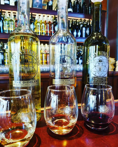 Three bottles of wine labeled "The Diddler," "Jane Pink," and "Tarzan Red" with two empty glasses in front.