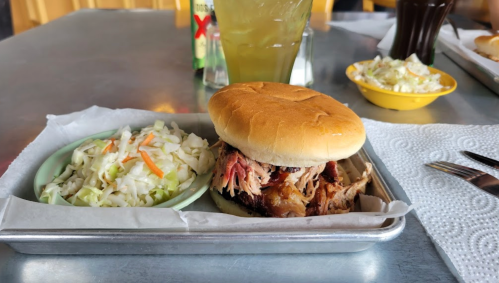 A pulled pork sandwich on a bun with a side of coleslaw, served on a metal tray with napkins and drinks in the background.