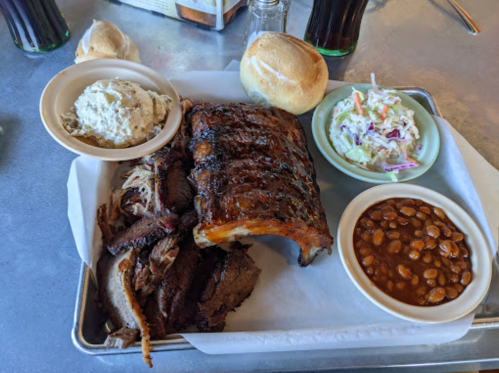 A platter of barbecue ribs, brisket, coleslaw, baked beans, and rolls on a metal tray.