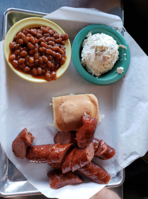 A plate with sliced sausage, a bun, baked beans, and coleslaw served in bowls.