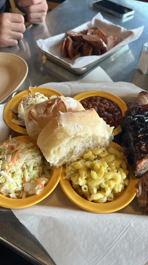 A plate with pulled pork, macaroni and cheese, coleslaw, baked beans, and two rolls, with a side of sausage in the background.