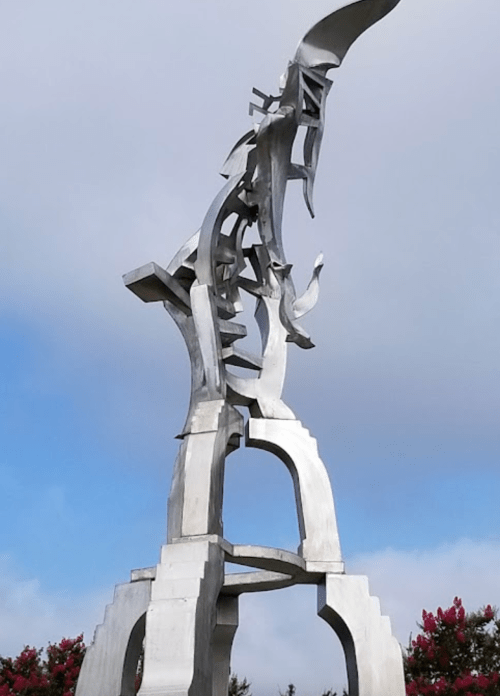 A tall, abstract metal sculpture with dynamic shapes, set against a cloudy sky and surrounded by greenery.