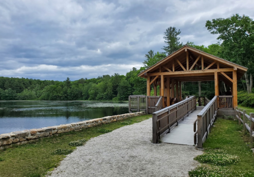 A wooden pavilion with a bridge overlooks a calm lake surrounded by lush greenery under a cloudy sky.