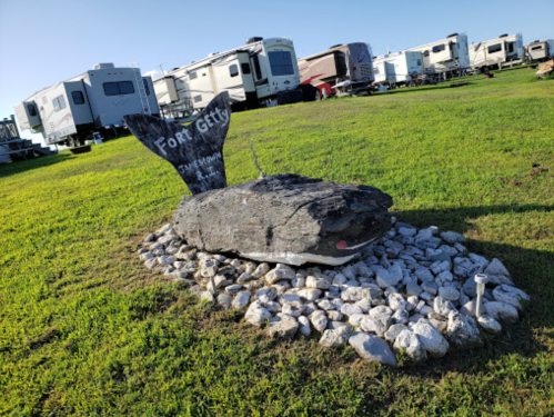 A wooden fish sculpture with a sign reading "Fort Getty" surrounded by rocks, set in a grassy area with RVs in the background.