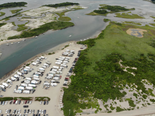 Aerial view of a river winding through green marshland, with a parking area for RVs and boats along the shore.