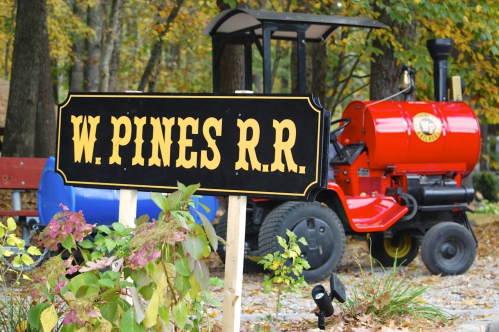 Sign for "W. Pines R.R." in front of a red miniature train engine, surrounded by autumn foliage.