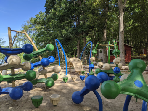 Colorful playground equipment with climbing structures and play areas surrounded by trees and a wooden cabin in the background.
