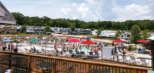A busy pool area with people relaxing, surrounded by RVs and umbrellas, under a partly cloudy sky.