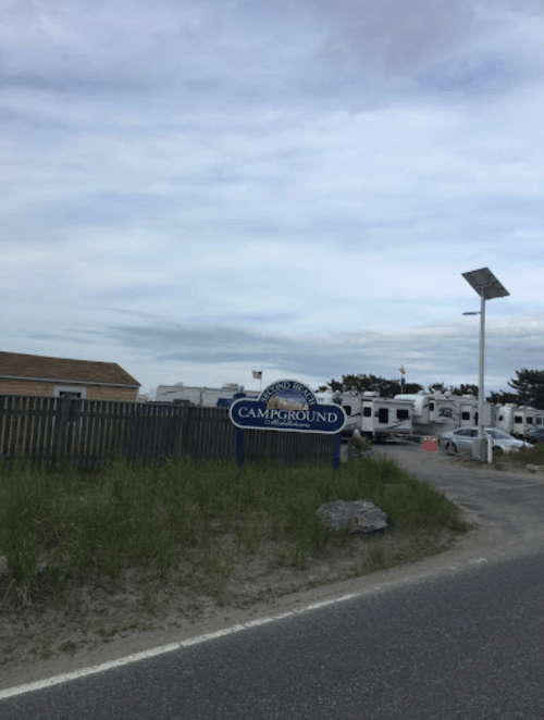 Sign for a campground near a road, with RVs parked in the background and a cloudy sky above.