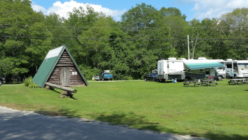 A grassy campsite with an A-frame cabin, RVs, and picnic tables surrounded by trees under a blue sky.