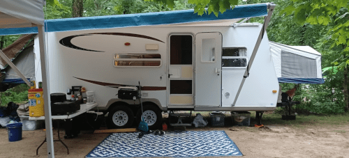 A white camper trailer parked in a wooded area, with a table set up outside and a blue rug on the ground.