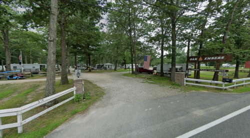 Entrance to Oak Embers Campground, featuring a sign, gravel road, and RVs surrounded by trees.