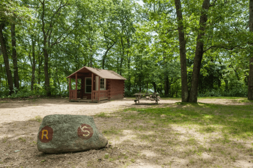 A small red cabin surrounded by trees, with a picnic table nearby and a large rock marked with "R" and "S."