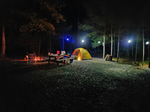 A campsite at night with a yellow tent, picnic table, and string lights illuminating the surrounding trees.