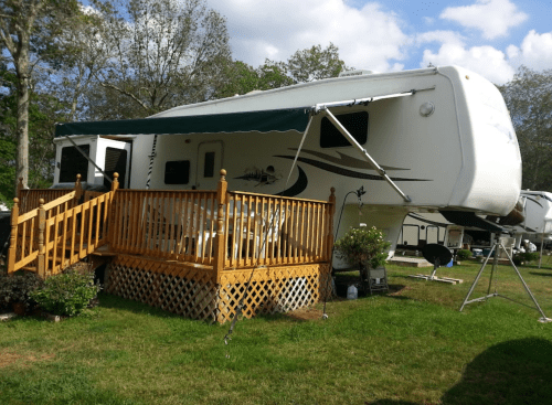A parked RV with a wooden deck and awning, surrounded by greenery and outdoor furniture.