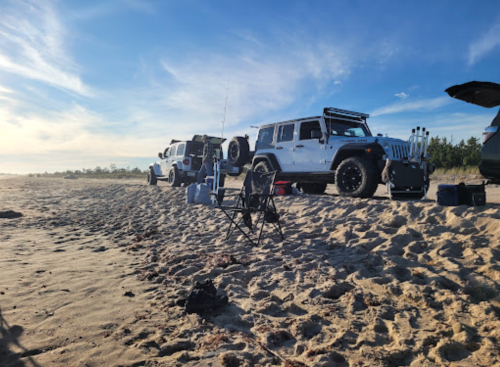 Two off-road vehicles parked on a sandy beach, with fishing gear and a chair set up nearby under a clear sky.