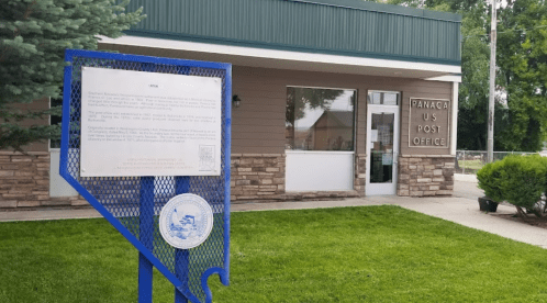 Sign in front of the Panaca U.S. Post Office, featuring a stone facade and green awning. Grass in foreground.