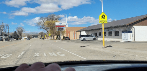 A view of a street with a market, a crosswalk sign, and a cloudy sky, seen from inside a vehicle.