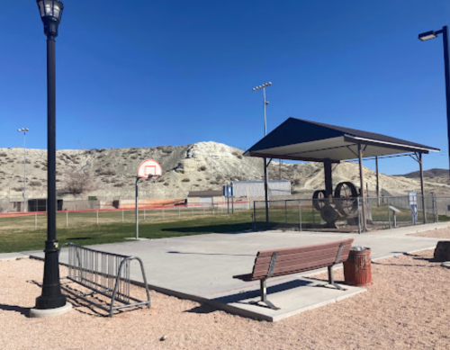 A basketball court with a bench and a shaded area, surrounded by a fence and hills in the background.