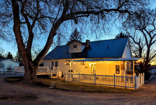 A charming white house with a blue roof, surrounded by trees and illuminated by string lights at dusk.