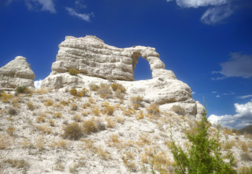 A rocky formation with a natural arch, surrounded by sparse vegetation under a blue sky with fluffy clouds.