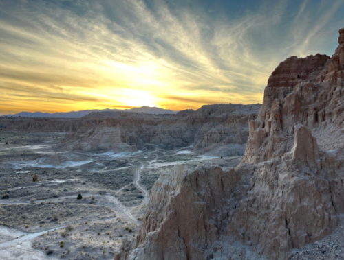 Sunset over rugged, layered rock formations in a vast desert landscape, with wispy clouds in the sky.