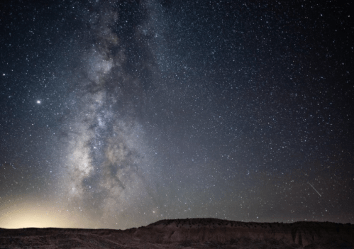 A stunning night sky filled with stars and the Milky Way, above a dark landscape with distant hills.