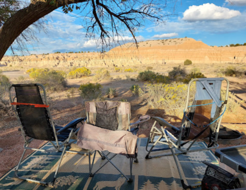 Two camping chairs sit on a rug, overlooking a desert landscape with hills and scattered vegetation under a blue sky.