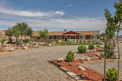 A red house with a tan roof surrounded by gravel, greenery, and rocks under a clear blue sky.