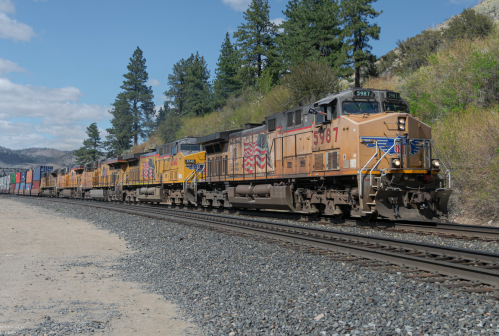 A freight train with multiple locomotives and colorful cargo containers travels along a rocky track surrounded by trees.