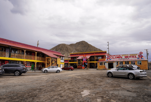 A colorful motel with clown-themed decor, surrounded by mountains and parked cars under a cloudy sky.