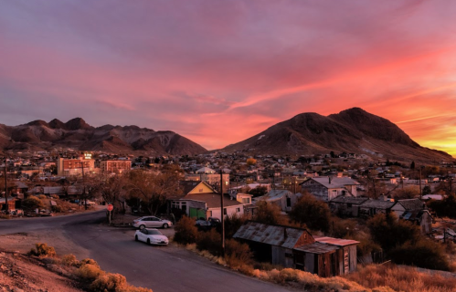 A sunset over a small town nestled between mountains, with houses and a winding road in the foreground.
