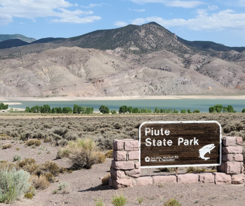 Sign for Piute State Park with mountains and a lake in the background under a clear blue sky.