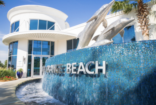 A modern building with a dolphin sculpture and a water feature, featuring the words "Orange Beach" prominently displayed.