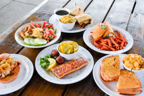 A variety of dishes on plates, including salmon, sandwiches, sweet potato fries, and salads, on a wooden table.