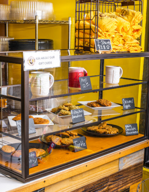 A display case filled with various baked goods and t-shirts, set against a bright yellow wall.