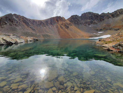 A serene mountain lake with clear water reflecting the rocky landscape and cloudy sky.