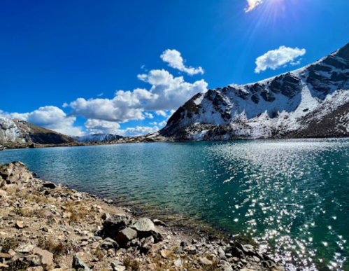 A serene mountain lake reflecting sunlight, surrounded by rocky shores and snow-capped peaks under a blue sky with clouds.