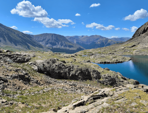 A scenic mountain landscape featuring rocky terrain, a calm lake, and blue skies with scattered clouds.