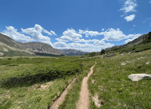 A winding dirt path leads through a lush green valley, surrounded by mountains and a bright blue sky with fluffy clouds.