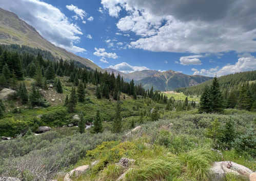 A scenic mountain landscape with lush greenery, a stream, and dramatic clouds in a blue sky.