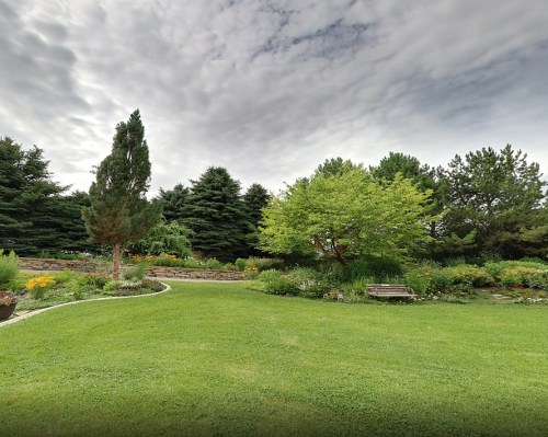 Lush garden with green grass, trees, and colorful flowers under a cloudy sky. A pathway winds through the landscape.