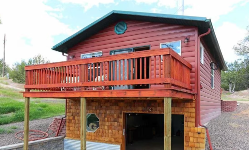 A wooden cabin with a balcony, set on stilts, surrounded by gravel and greenery under a blue sky.