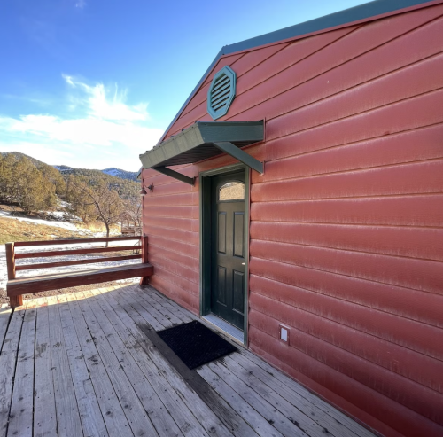 A rustic red cabin with a green door and wooden deck, set against a mountainous landscape under a clear blue sky.