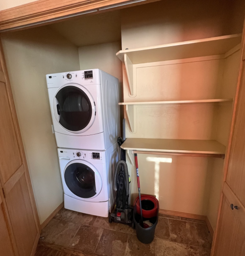 A laundry closet featuring stacked white washer and dryer units, with shelves and cleaning supplies below.