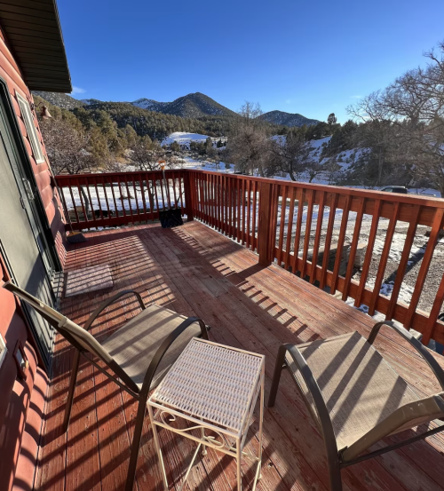 A wooden deck with two chairs and a small table, overlooking a snowy landscape and mountains in the distance.